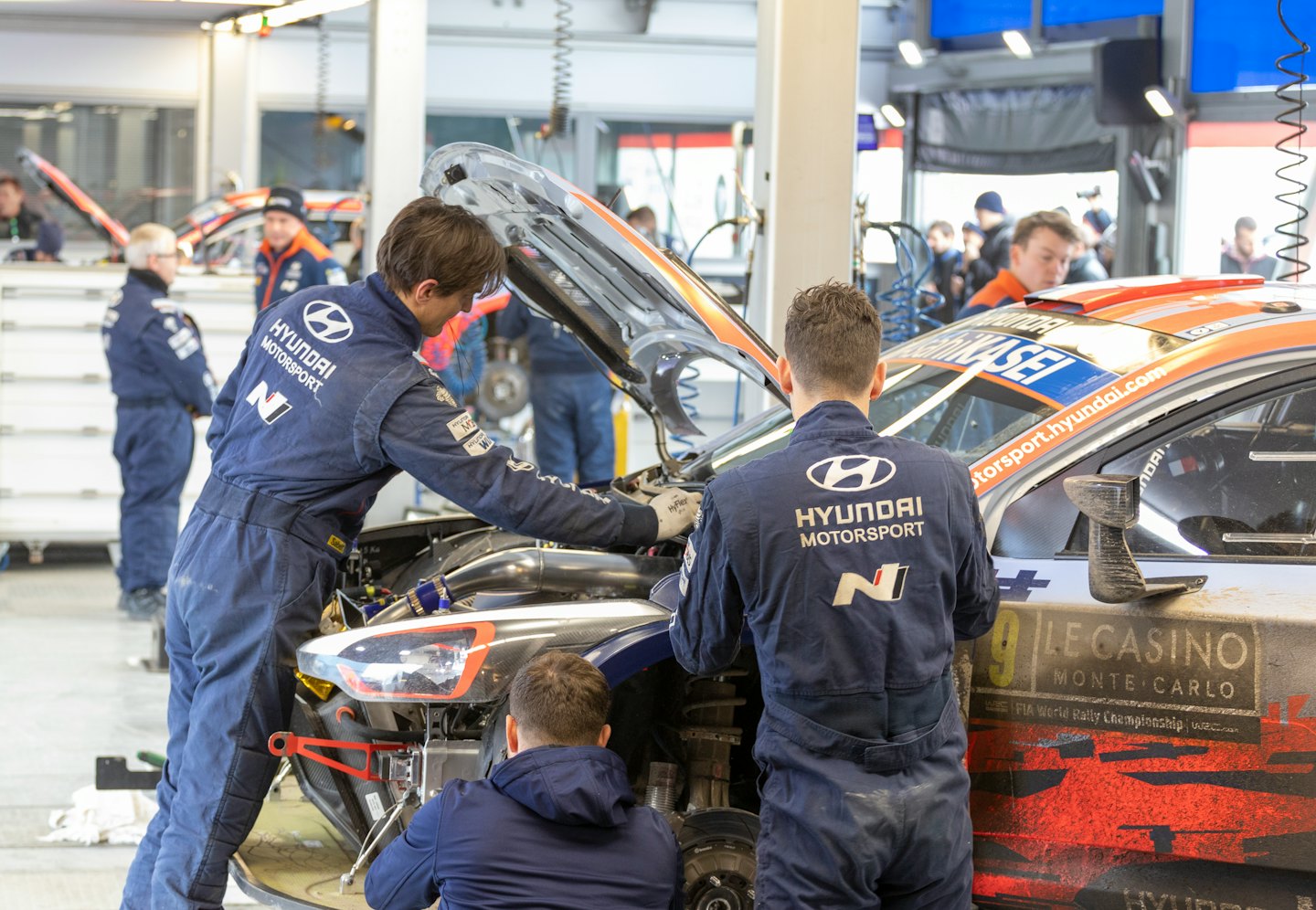 Meccanici in tuta blu lavorano su un'auto da rally con il cofano aperto.